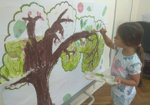 Zdjęcie przedstawia dziewczynkę stojącą przy dużym plakacie drzewa, która je maluje jego elementy farbami